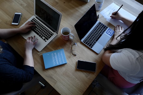 A wooden table supports two laptops, each operated by different individuals. A pair of reading glasses and a blue book titled 'Empowered Women Empower Women' rest between them. Smartphones, notebooks, and coffee mugs are also present, contributing to a productive workspace setting.
