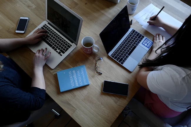 A wooden table supports two laptops, each operated by different individuals. A pair of reading glasses and a blue book titled 'Empowered Women Empower Women' rest between them. Smartphones, notebooks, and coffee mugs are also present, contributing to a productive workspace setting.