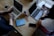 A wooden table supports two laptops, each operated by different individuals. A pair of reading glasses and a blue book titled 'Empowered Women Empower Women' rest between them. Smartphones, notebooks, and coffee mugs are also present, contributing to a productive workspace setting.