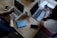 A wooden table supports two laptops, each operated by different individuals. A pair of reading glasses and a blue book titled 'Empowered Women Empower Women' rest between them. Smartphones, notebooks, and coffee mugs are also present, contributing to a productive workspace setting.