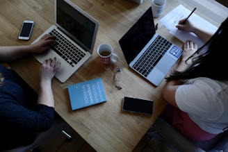 A wooden table supports two laptops, each operated by different individuals. A pair of reading glasses and a blue book titled 'Empowered Women Empower Women' rest between them. Smartphones, notebooks, and coffee mugs are also present, contributing to a productive workspace setting.