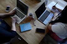 A wooden table supports two laptops, each operated by different individuals. A pair of reading glasses and a blue book titled 'Empowered Women Empower Women' rest between them. Smartphones, notebooks, and coffee mugs are also present, contributing to a productive workspace setting.