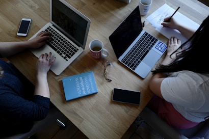 A wooden table supports two laptops, each operated by different individuals. A pair of reading glasses and a blue book titled 'Empowered Women Empower Women' rest between them. Smartphones, notebooks, and coffee mugs are also present, contributing to a productive workspace setting.
