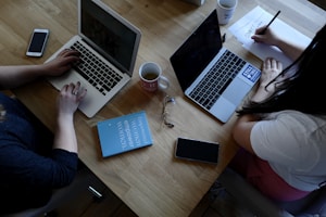 A wooden table supports two laptops, each operated by different individuals. A pair of reading glasses and a blue book titled 'Empowered Women Empower Women' rest between them. Smartphones, notebooks, and coffee mugs are also present, contributing to a productive workspace setting.