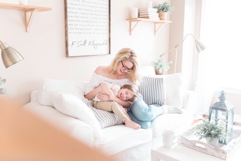 A cozy living room setting with a woman and child sitting together on a white couch. The woman, with blond hair and glasses, is embracing the child, who is smiling. The room is decorated with a framed quote, a couple of striped cushions, and a potted plant on a wooden shelf. The bright space features soft lighting and white decor, including a tray with greenery and a lantern on a coffee table.