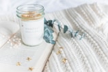 A scented candle in a glass jar with the label 'Manifest and Chill' sits on an open book with a textured knit blanket in the background. Sprigs of eucalyptus and small white flowers are placed nearby, along with scattered gold star confetti.
