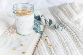 A scented candle in a glass jar with the label 'Manifest and Chill' sits on an open book with a textured knit blanket in the background. Sprigs of eucalyptus and small white flowers are placed nearby, along with scattered gold star confetti.