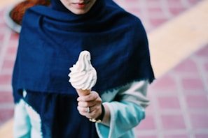 A person is holding a vanilla soft serve ice cream cone. They are wearing a dark blue shawl over a light-colored outfit. The background features a brick-patterned ground with a yellow stripe.