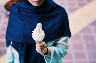 A person is holding a vanilla soft serve ice cream cone. They are wearing a dark blue shawl over a light-colored outfit. The background features a brick-patterned ground with a yellow stripe.