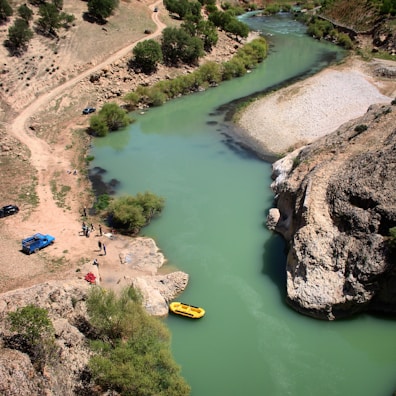 An aerial view of a winding turquoise river bordered by rocky and grassy terrain. A yellow inflatable boat is moored at the riverbank where several people gather near a blue truck on a dirt path. Sparse trees dot the landscape.