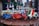A collection of small, colorful vintage toy vehicles, including scooters, cars, and vans, displayed on a wooden surface. The toys are intricately detailed and painted in bright colors, with a background of blurred decorative elements, suggesting a festive atmosphere.