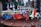 A collection of small, colorful vintage toy vehicles, including scooters, cars, and vans, displayed on a wooden surface. The toys are intricately detailed and painted in bright colors, with a background of blurred decorative elements, suggesting a festive atmosphere.