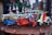 A collection of small, colorful vintage toy vehicles, including scooters, cars, and vans, displayed on a wooden surface. The toys are intricately detailed and painted in bright colors, with a background of blurred decorative elements, suggesting a festive atmosphere.