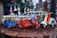 A collection of small, colorful vintage toy vehicles, including scooters, cars, and vans, displayed on a wooden surface. The toys are intricately detailed and painted in bright colors, with a background of blurred decorative elements, suggesting a festive atmosphere.