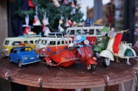 A collection of small, colorful vintage toy vehicles, including scooters, cars, and vans, displayed on a wooden surface. The toys are intricately detailed and painted in bright colors, with a background of blurred decorative elements, suggesting a festive atmosphere.