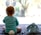 A baby wearing light blue pajamas with a fluffy white tail sits on a soft carpet facing a large window. The baby's head is adorned with soft bunny ears. Beside the baby sits a plush blue bunny toy, mirroring the baby's attire and ears.