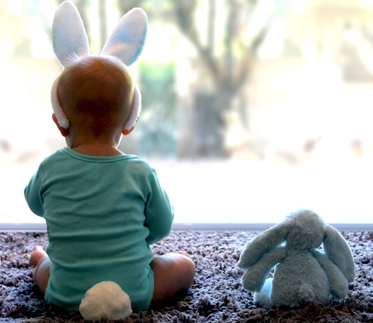 A baby wearing light blue pajamas with a fluffy white tail sits on a soft carpet facing a large window. The baby's head is adorned with soft bunny ears. Beside the baby sits a plush blue bunny toy, mirroring the baby's attire and ears.