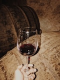 A hand holds a glass of red wine against the backdrop of large wooden barrels, presumably in a dimly lit wine cellar. The ambiance is rustic, with a warm, earthy tone.