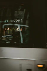 A blurry reflection of a shop window with mannequins displaying clothing is partially obscured by the passing of a large vehicle. The words 'MOODS' can be seen, suggesting a storefront or advertisement. The scene is set in an urban environment with a dark and slightly mysterious ambiance caused by the lighting and motion blur.