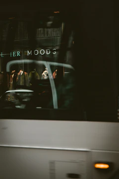 A blurry reflection of a shop window with mannequins displaying clothing is partially obscured by the passing of a large vehicle. The words 'MOODS' can be seen, suggesting a storefront or advertisement. The scene is set in an urban environment with a dark and slightly mysterious ambiance caused by the lighting and motion blur.
