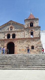 A rustic stone church with a prominent bell tower and weathered brick accents. Several people are standing on the wide stone steps leading to the arched entrance, which gives a sense of a casual gathering. The church features a cross and a traditional tiled roof. The sky is clear and blue, adding to the serene atmosphere.