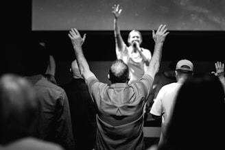 A black and white photo of a crowd facing forward with hands raised, likely at a concert or religious gathering. In the background, a person is on stage holding a microphone and engaging with the audience.