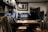 A cozy coffee shop interior featuring a wooden counter with stools, a variety of brewing equipment, and a person working behind the bar. Shelves filled with jars, cups, and bottles line the walls, and a framed black-and-white photograph hangs above the espresso machine.