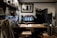 A cozy coffee shop interior featuring a wooden counter with stools, a variety of brewing equipment, and a person working behind the bar. Shelves filled with jars, cups, and bottles line the walls, and a framed black-and-white photograph hangs above the espresso machine.