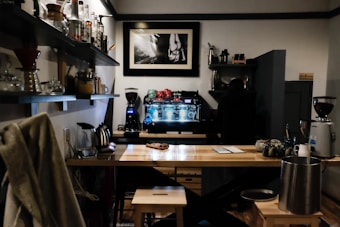 A cozy coffee shop interior featuring a wooden counter with stools, a variety of brewing equipment, and a person working behind the bar. Shelves filled with jars, cups, and bottles line the walls, and a framed black-and-white photograph hangs above the espresso machine.