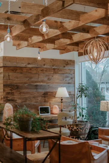 A cozy interior with wooden decor, including a ceiling with exposed wooden beams and walls. The space is furnished with a wooden table, chairs, and a desk setup featuring a laptop and a salt lamp. Potted plants add greenery, while soft lighting from hanging bulbs and a floor lamp creates a warm ambiance. The large window provides natural light and a view of the outside trees.
