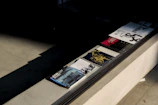 Several magazines or books are neatly arranged on a ledge next to a large window. The publications have various covers, featuring artistic images and different colors, with one prominently displaying the text 'ARTMAP'. The light casts distinct shadows across the textured surface.