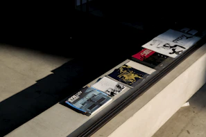 Several magazines or books are neatly arranged on a ledge next to a large window. The publications have various covers, featuring artistic images and different colors, with one prominently displaying the text 'ARTMAP'. The light casts distinct shadows across the textured surface.
