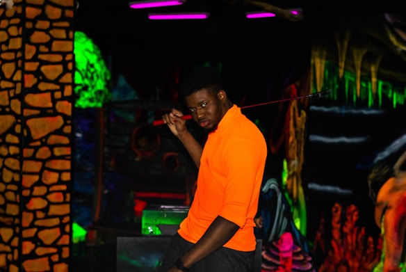 A person wearing a bright orange shirt holds a golf club in a dark, neon-lit environment that resembles a cosmic or underworld-themed setting. The background features vibrant colors and abstract patterns, with elements resembling glowing stones and caverns.