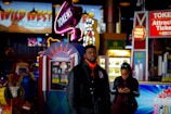 A bustling arcade environment with neon signs, colorful game machines, and a man in a black jacket standing in the foreground. A woman is seen in the background using her phone. Bright signage advertising tokens and attractions adds to the vibrant atmosphere.