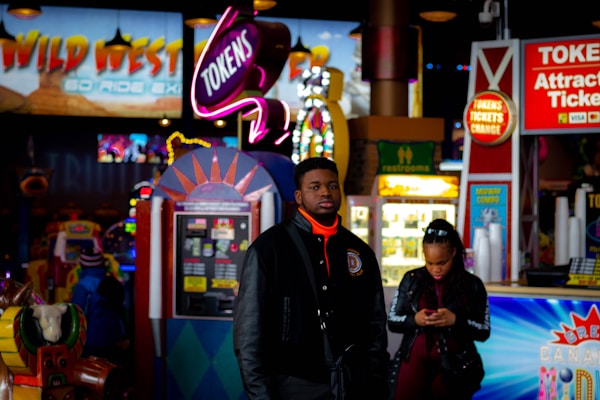 A bustling arcade environment with neon signs, colorful game machines, and a man in a black jacket standing in the foreground. A woman is seen in the background using her phone. Bright signage advertising tokens and attractions adds to the vibrant atmosphere.