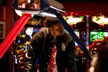 A man stands in an arcade, with vibrant gaming machines glowing in the background. He wears a black jacket with a colorful shirt underneath. The scene is brightly lit with neon lights adding to the playful ambiance.