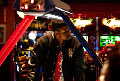 A man stands in an arcade, with vibrant gaming machines glowing in the background. He wears a black jacket with a colorful shirt underneath. The scene is brightly lit with neon lights adding to the playful ambiance.