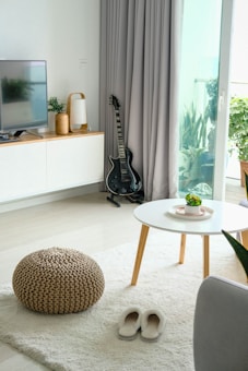A cozy living room with a modern aesthetic featuring a guitar in the corner beside floor-length gray curtains. A television is placed on a wooden cabinet along with a small potted plant and a decorative lamp. The white coffee table with wooden legs holds a small succulent plant. A knitted pouf and a pair of soft slippers rest on a plush area rug.