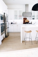 A modern kitchen with white cabinetry and stainless steel appliances. Two wooden stools with metal legs are positioned by a white island. The floor is made of light wood, and there are decorative items such as cutting boards and a wine bottle on the countertop. A large ceiling light fixture hangs overhead.