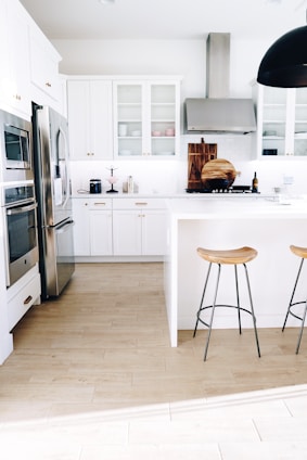 A modern kitchen with white cabinetry and stainless steel appliances. Two wooden stools with metal legs are positioned by a white island. The floor is made of light wood, and there are decorative items such as cutting boards and a wine bottle on the countertop. A large ceiling light fixture hangs overhead.
