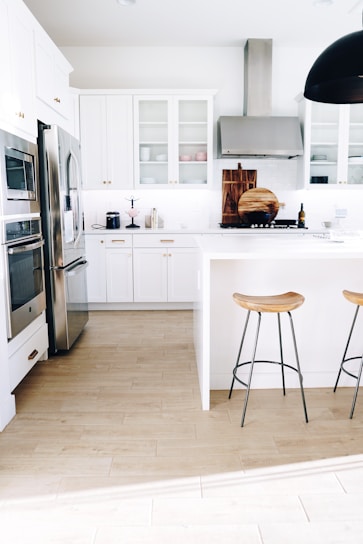 A modern kitchen with white cabinetry and stainless steel appliances. Two wooden stools with metal legs are positioned by a white island. The floor is made of light wood, and there are decorative items such as cutting boards and a wine bottle on the countertop. A large ceiling light fixture hangs overhead.