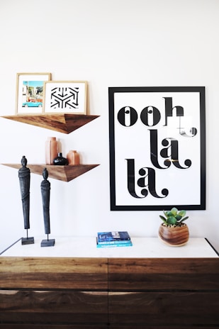A modern interior setup features a wooden console with two standing statues on top. Above, a geometric wooden shelf holds two framed artworks - one abstract and another colorful landscape. Beside the shelf, a large framed print displays the phrase 'ooh la la'. A small stack of books and a potted succulent plant sit on the console, adding a touch of color and greenery to the setting.