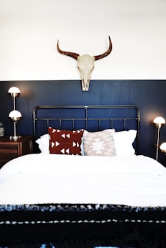 A stylish bedroom features a dark metal bed frame against a two-toned wall with a white upper section and dark blue lower section. Above the bed, a decorative skull with horns is mounted on the wall. There are two patterned throw pillows in red and white on the neatly made bed with white linens. On either side of the bed are wooden nightstands with modern spherical lamps.