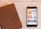A close-up view of a smartphone displaying various podcast covers on its screen, alongside a brown leather-covered notebook lying on a table. In the background, part of a white keyboard and another notebook with a checked pattern are visible.