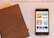 A close-up view of a smartphone displaying various podcast covers on its screen, alongside a brown leather-covered notebook lying on a table. In the background, part of a white keyboard and another notebook with a checked pattern are visible.