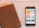 A close-up view of a smartphone displaying various podcast covers on its screen, alongside a brown leather-covered notebook lying on a table. In the background, part of a white keyboard and another notebook with a checked pattern are visible.