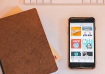 A close-up view of a smartphone displaying various podcast covers on its screen, alongside a brown leather-covered notebook lying on a table. In the background, part of a white keyboard and another notebook with a checked pattern are visible.
