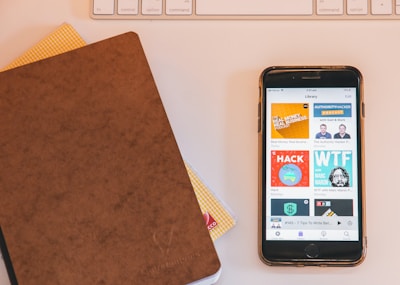 A close-up view of a smartphone displaying various podcast covers on its screen, alongside a brown leather-covered notebook lying on a table. In the background, part of a white keyboard and another notebook with a checked pattern are visible.