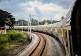 A long train with multiple carriages curves along a railway track surrounded by lush greenery. The sky is partially cloudy, creating a serene backdrop for the countryside scene. The train appears luxurious and polished, reflecting light off its metallic surface.