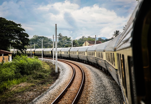 A long train with multiple carriages curves along a railway track surrounded by lush greenery. The sky is partially cloudy, creating a serene backdrop for the countryside scene. The train appears luxurious and polished, reflecting light off its metallic surface.